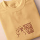 A yellow crew neck short sleeve t-shirt, with an embroidered brown thread design of a cute singing cat wearing a cowboy hat and the song lyric country roads, take me home to the place I belong