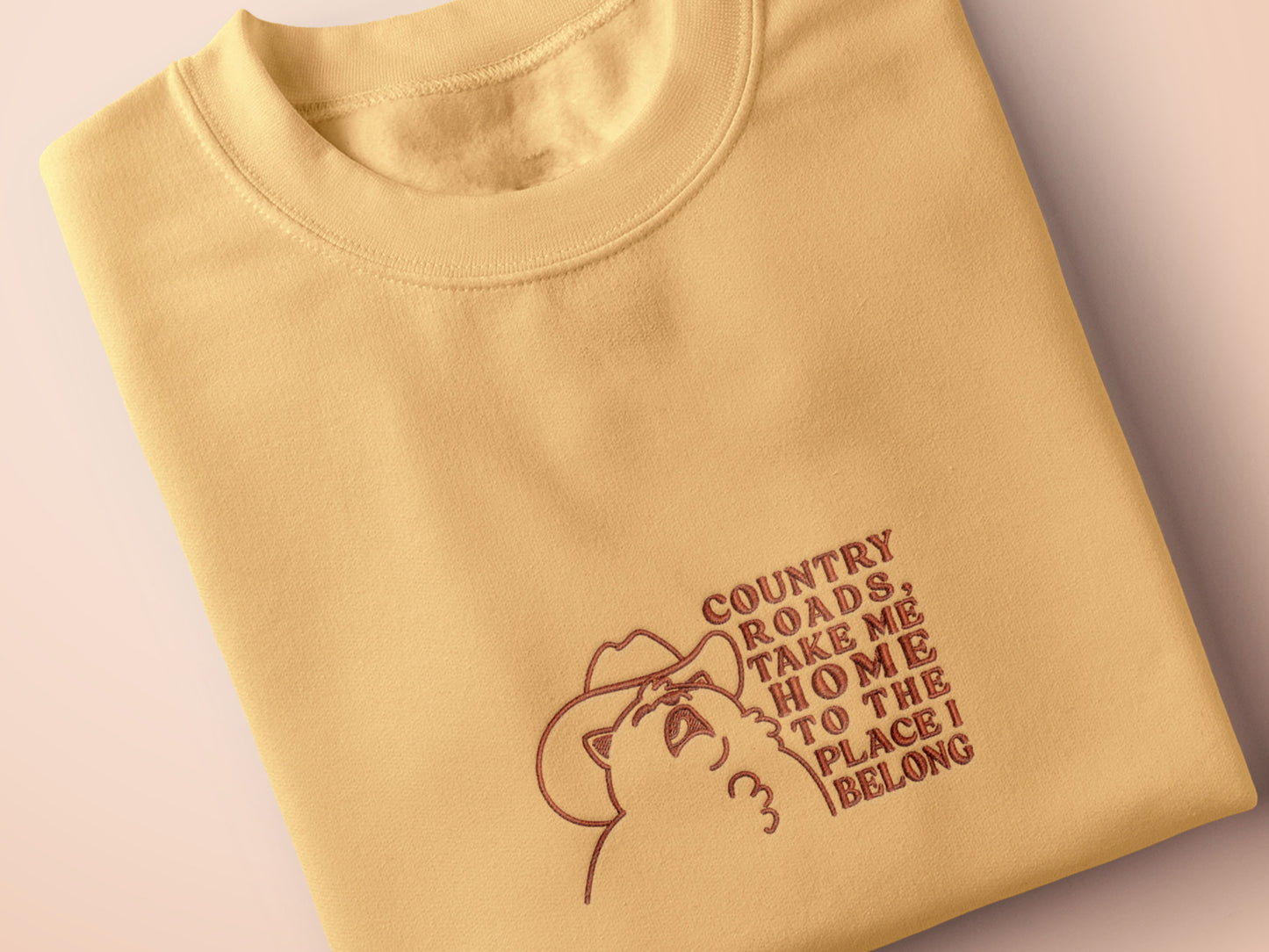 A yellow crew neck short sleeve t-shirt, with an embroidered brown thread design of a cute singing cat wearing a cowboy hat and the song lyric country roads, take me home to the place I belong