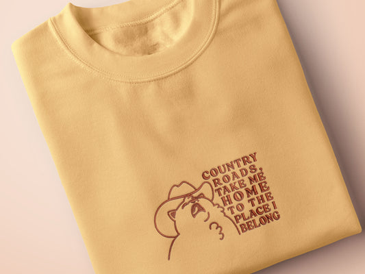 A yellow crew neck short sleeve t-shirt, with an embroidered brown thread design of a cute singing cat wearing a cowboy hat and the song lyric country roads, take me home to the place I belong