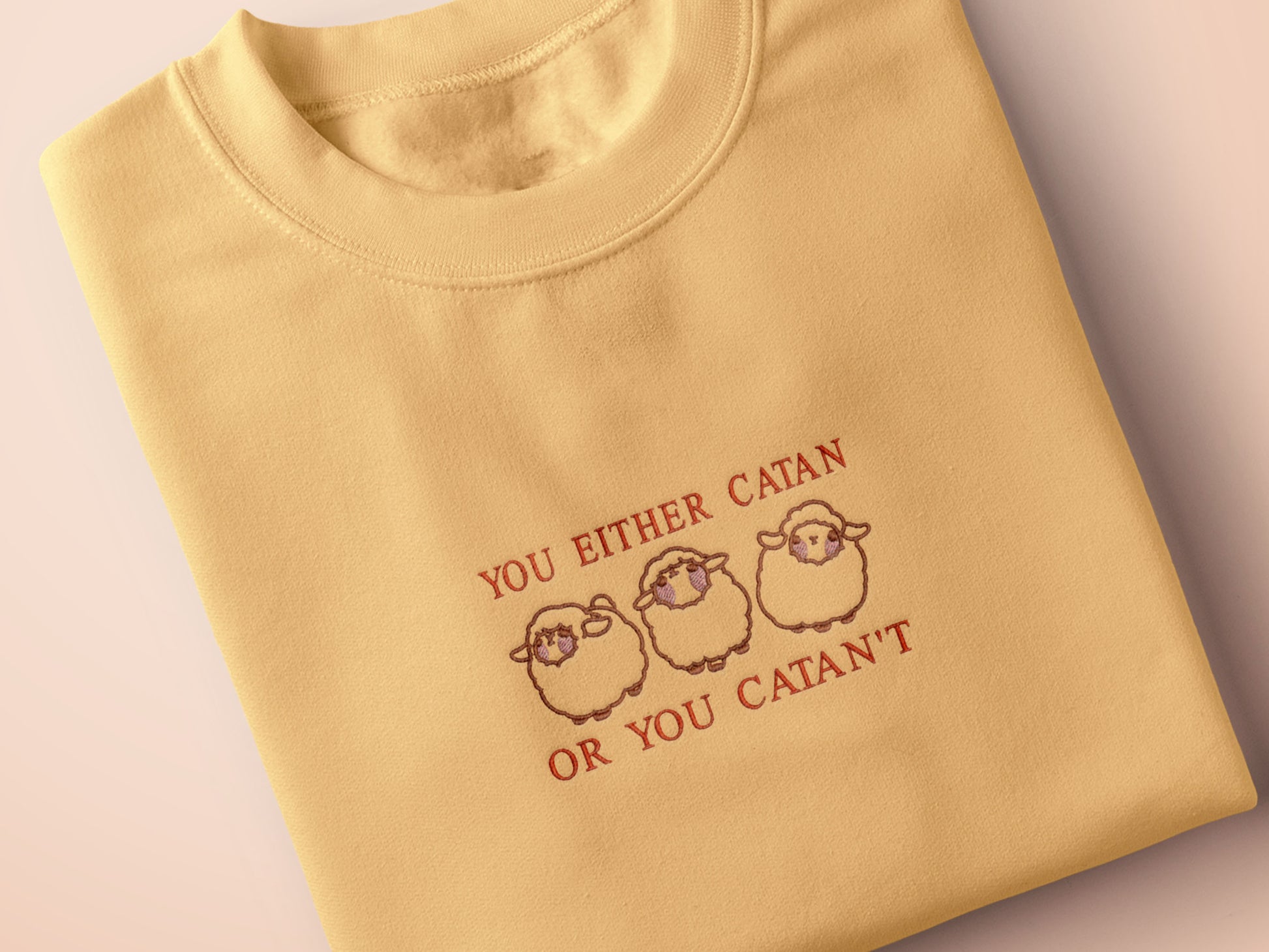 A yellow top with a brown and red embroidered design of cute chibi sheep with the text you either catan or you catan't