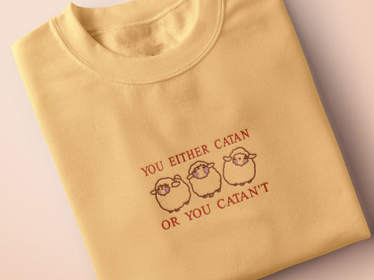 A yellow top with a brown and red embroidered design of cute chibi sheep with the text you either catan or you catan't