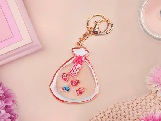 A large transparent acrylic shaker keychain with gold keyring, filled with mini chibi shrimps and labelled as emotional support shrimp