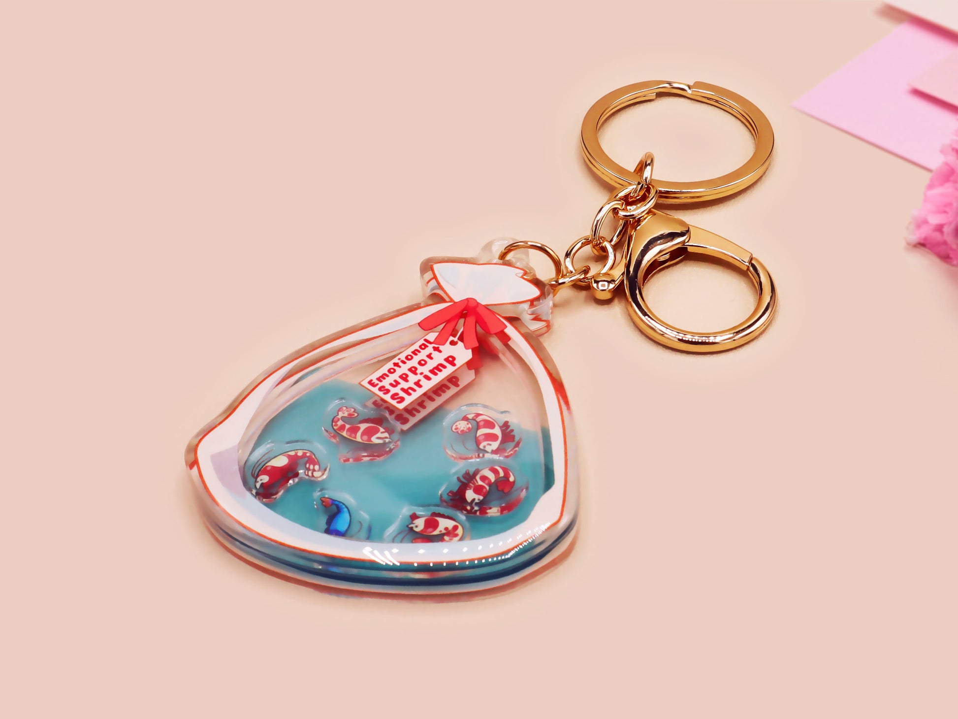 A large transparent acrylic shaker keychain with gold keyring, filled with mini chibi shrimps and labelled as emotional support shrimp