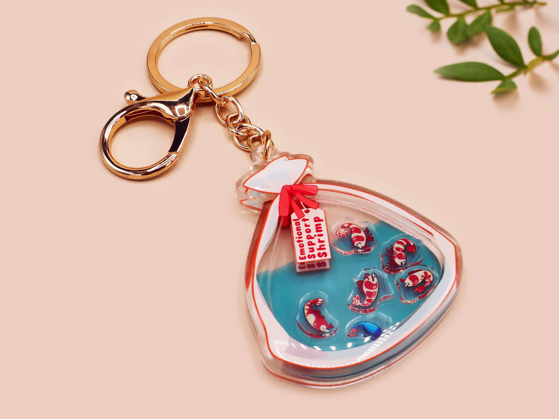 A large transparent acrylic shaker keychain with gold keyring, filled with mini chibi shrimps and labelled as emotional support shrimp