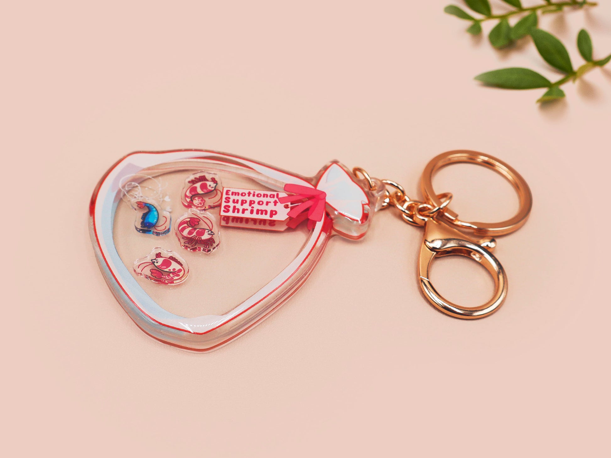 A large transparent acrylic shaker keychain with gold keyring, filled with mini chibi shrimps and labelled as emotional support shrimp