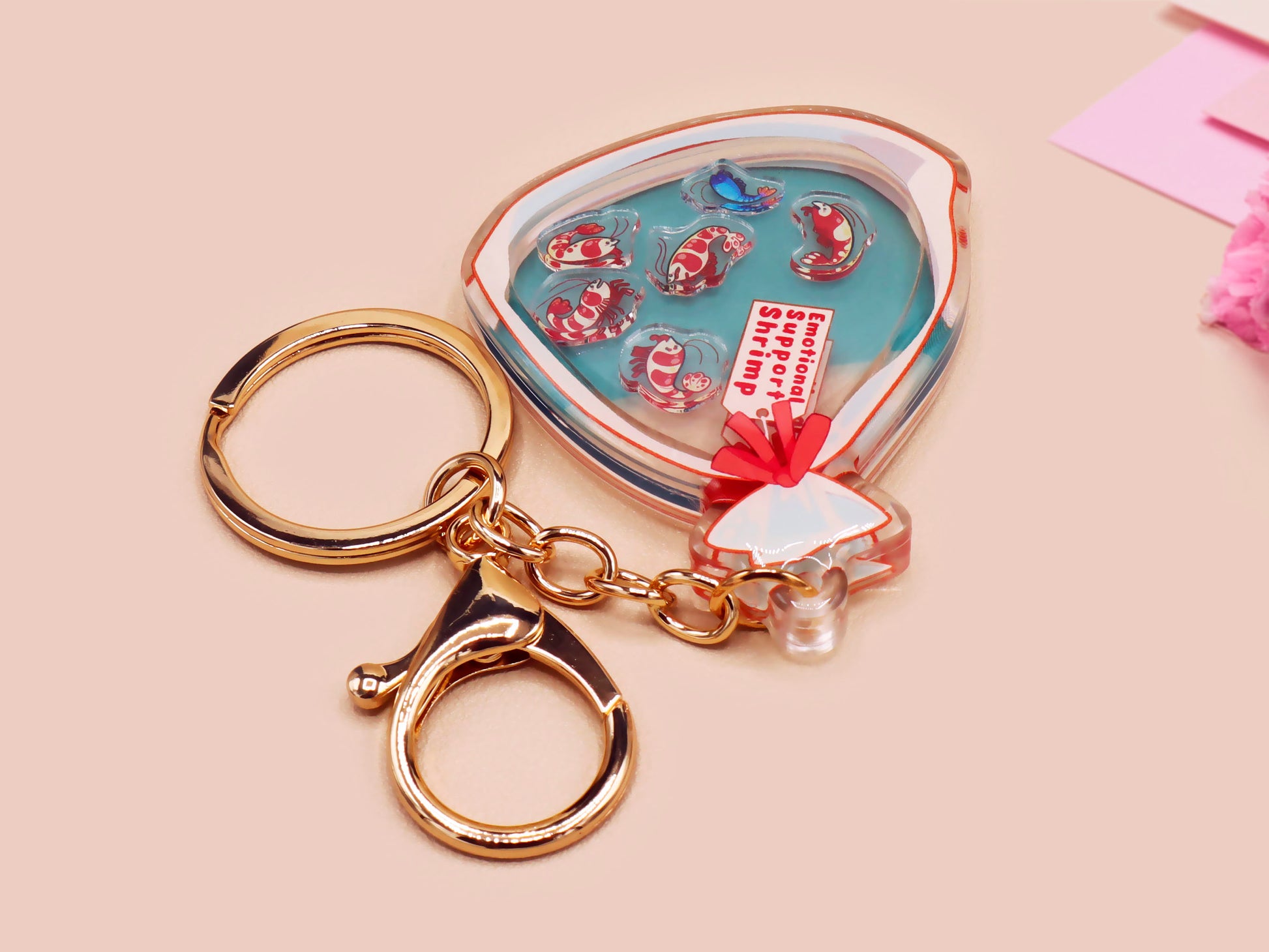 A large transparent acrylic shaker keychain with gold keyring, filled with mini chibi shrimps and labelled as emotional support shrimp