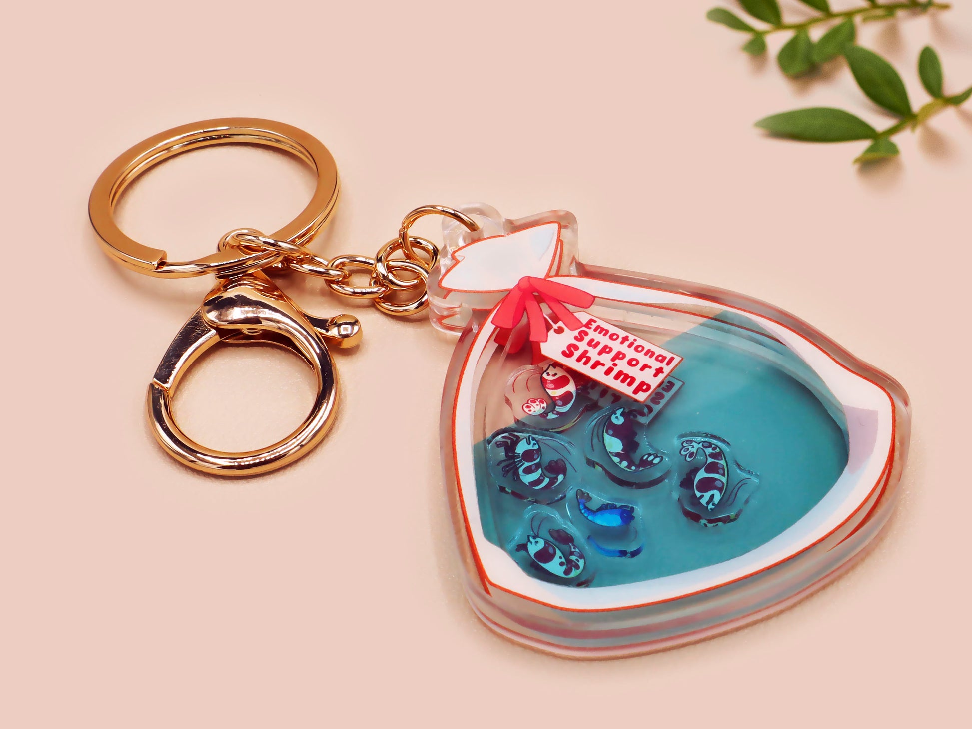 A large transparent acrylic shaker keychain with gold keyring, filled with mini chibi shrimps and labelled as emotional support shrimp