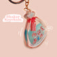 A large transparent acrylic shaker keychain with gold keyring, filled with mini chibi shrimps and labelled as emotional support shrimp