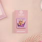 A clear acrylic pin badge with a pink heart-shaped pin back clasp. The design of the pin badge is of a cute chibi purple raccoon sat ona sofa wrapped in a blanket crying with the quote even baddies get saddies.
