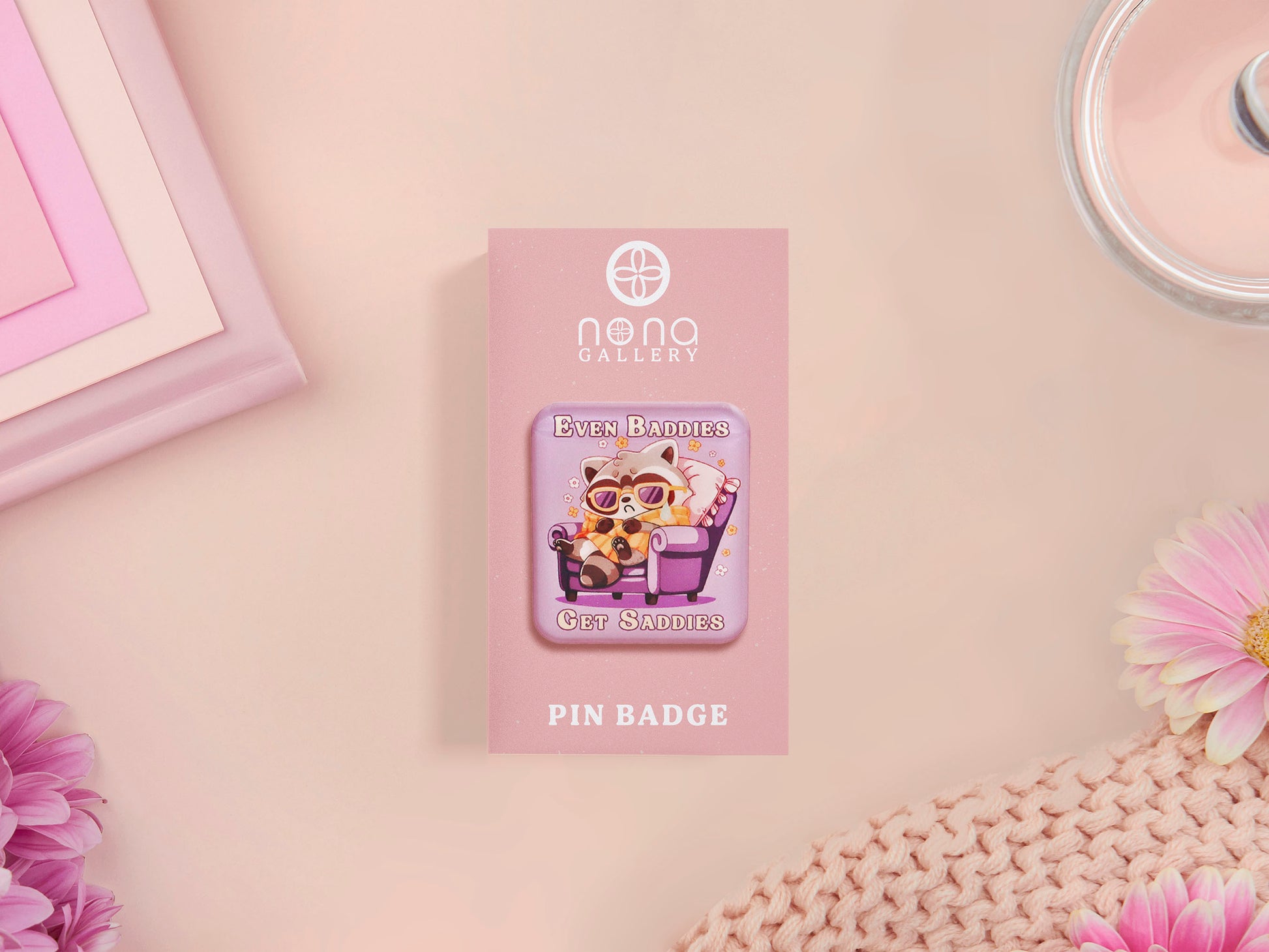 A clear acrylic pin badge with a pink heart-shaped pin back clasp. The design of the pin badge is of a cute chibi purple raccoon sat ona sofa wrapped in a blanket crying with the quote even baddies get saddies.