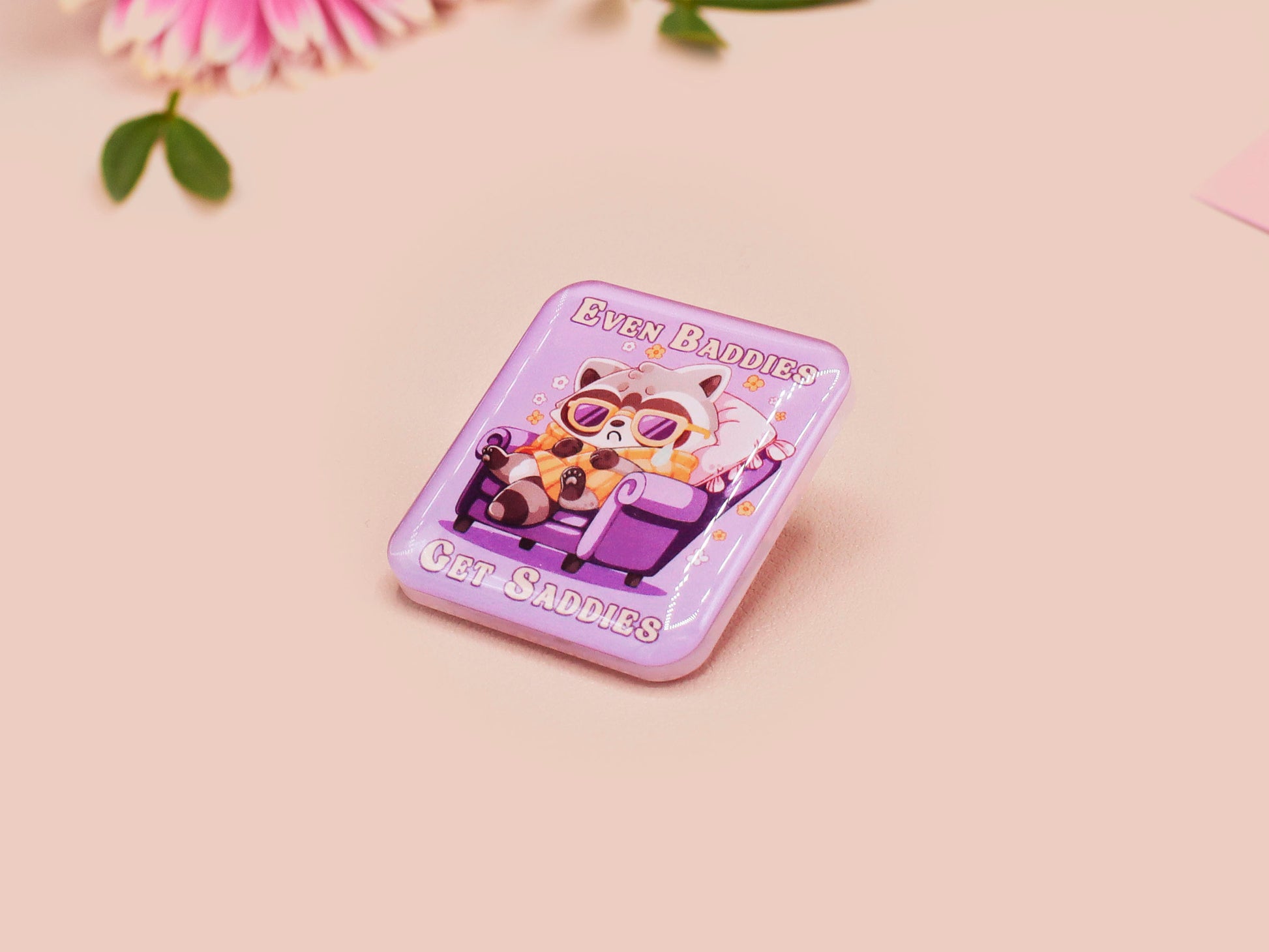 A clear acrylic pin badge with a pink heart-shaped pin back clasp. The design of the pin badge is of a cute chibi purple raccoon sat ona sofa wrapped in a blanket crying with the quote even baddies get saddies.
