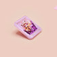A clear acrylic pin badge with a pink heart-shaped pin back clasp. The design of the pin badge is of a cute chibi purple raccoon sat ona sofa wrapped in a blanket crying with the quote even baddies get saddies.