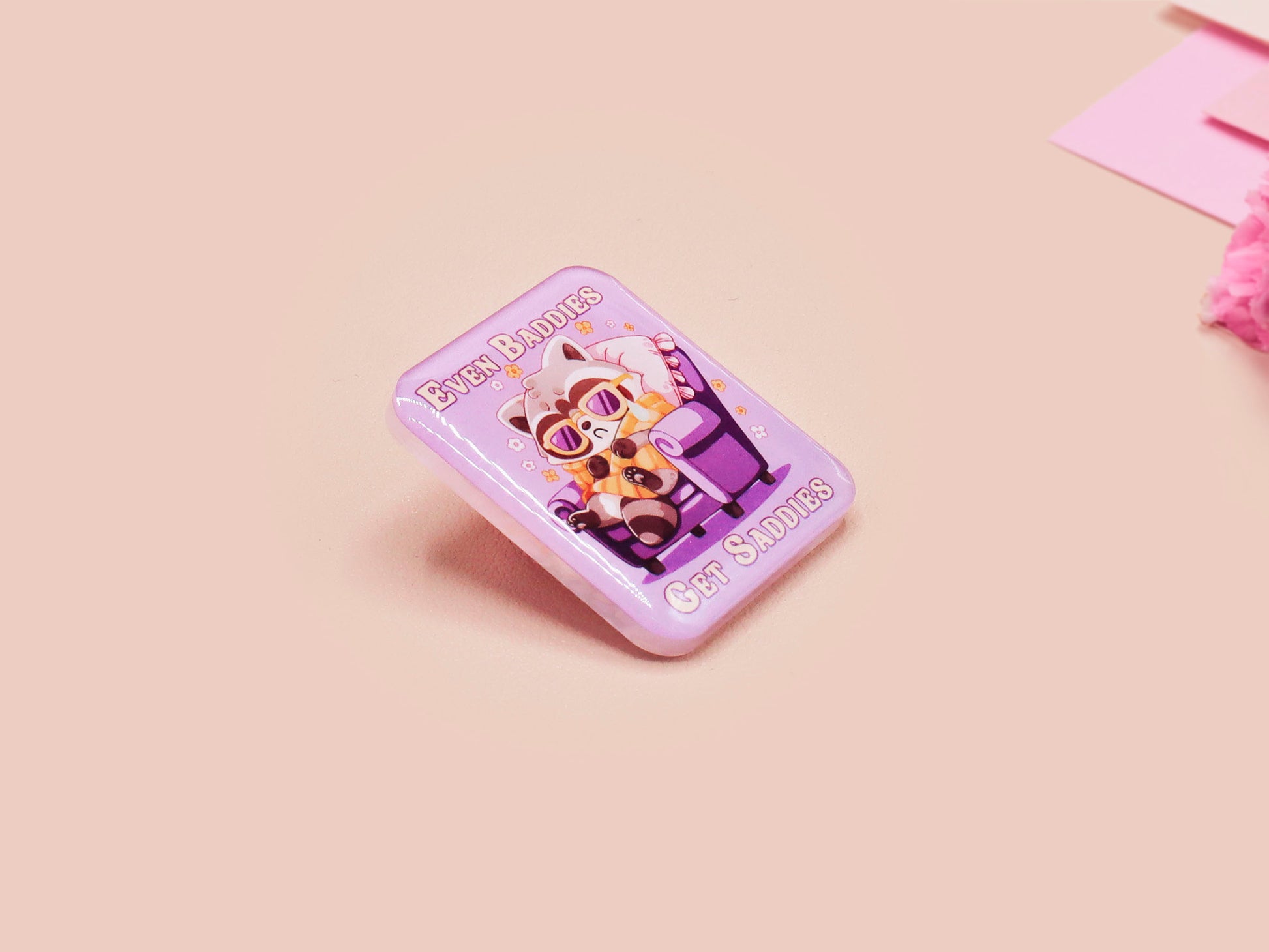 A clear acrylic pin badge with a pink heart-shaped pin back clasp. The design of the pin badge is of a cute chibi purple raccoon sat ona sofa wrapped in a blanket crying with the quote even baddies get saddies.