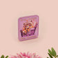 A clear acrylic pin badge with a pink heart-shaped pin back clasp. The design of the pin badge is of a cute chibi purple raccoon sat ona sofa wrapped in a blanket crying with the quote even baddies get saddies.