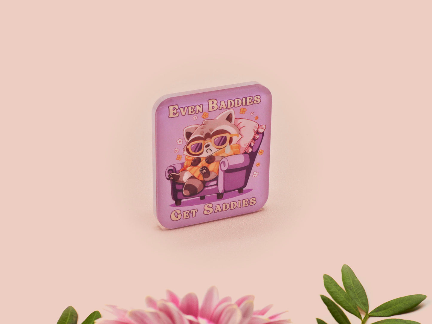 A clear acrylic pin badge with a pink heart-shaped pin back clasp. The design of the pin badge is of a cute chibi purple raccoon sat ona sofa wrapped in a blanket crying with the quote even baddies get saddies.