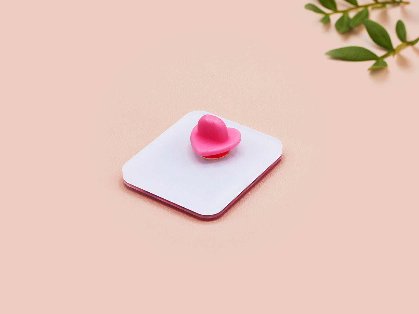 A clear acrylic pin badge with a pink heart-shaped pin back clasp. The design of the pin badge is of a cute chibi purple raccoon sat ona sofa wrapped in a blanket crying with the quote even baddies get saddies.