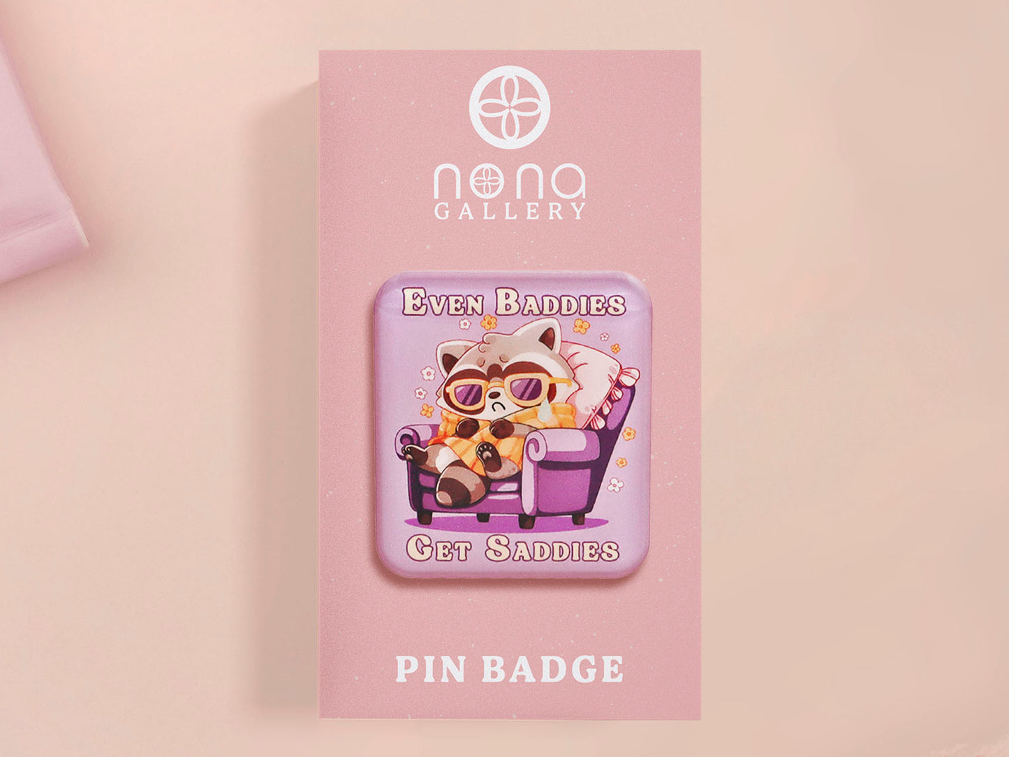 A clear acrylic pin badge with a pink heart-shaped pin back clasp. The design of the pin badge is of a cute chibi purple raccoon sat ona sofa wrapped in a blanket crying with the quote even baddies get saddies.
