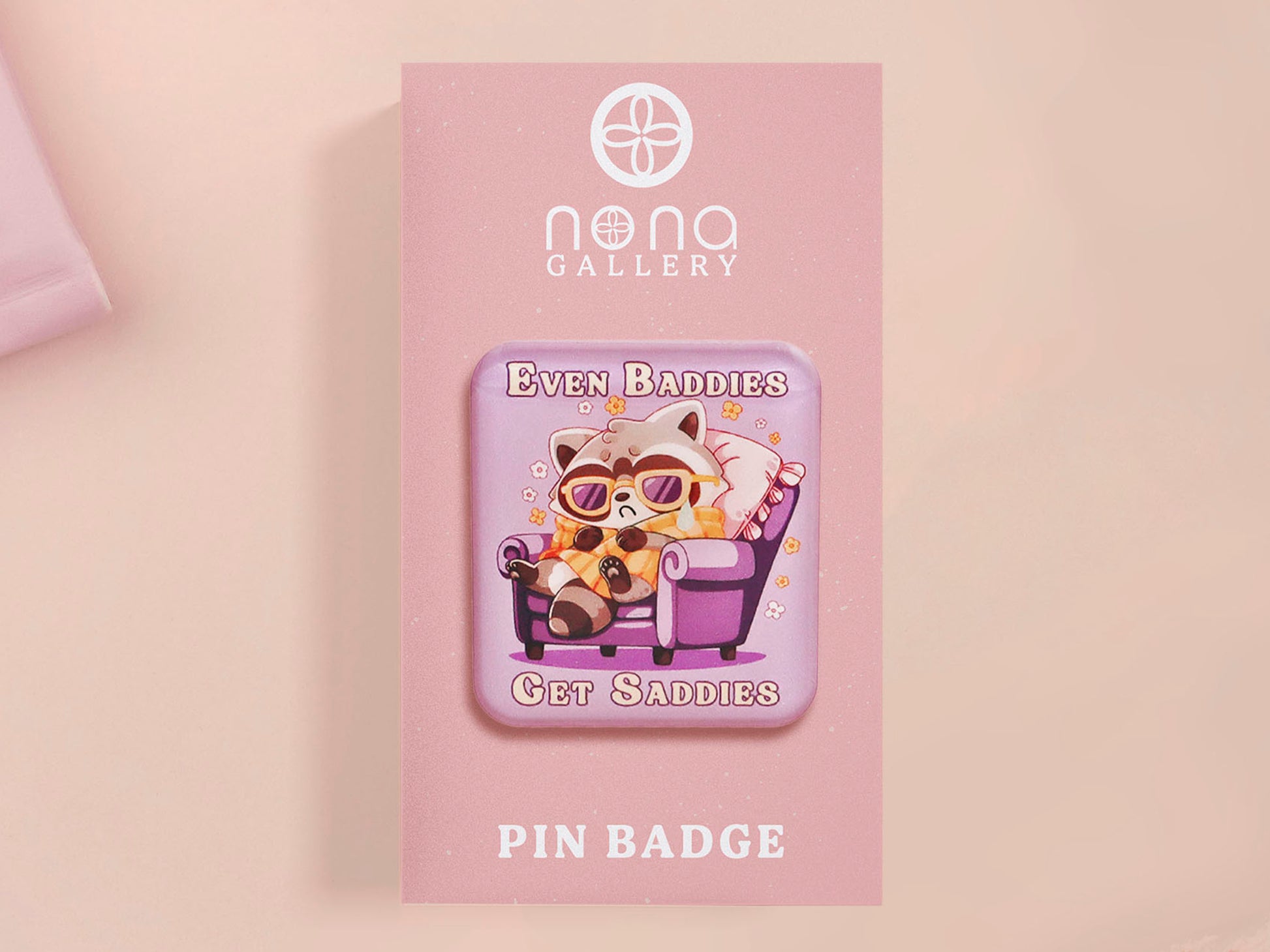 A clear acrylic pin badge with a pink heart-shaped pin back clasp. The design of the pin badge is of a cute chibi purple raccoon sat ona sofa wrapped in a blanket crying with the quote even baddies get saddies.