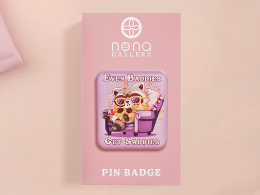 A clear acrylic pin badge with a pink heart-shaped pin back clasp. The design of the pin badge is of a cute chibi purple raccoon sat ona sofa wrapped in a blanket crying with the quote even baddies get saddies.