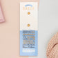 The back of the long white and blue backing card with gold accents. The backing card details the symbolism of the different design elements of the Baker's Talisman pin. Also shown is the two gold locking pin backs attached to the card.