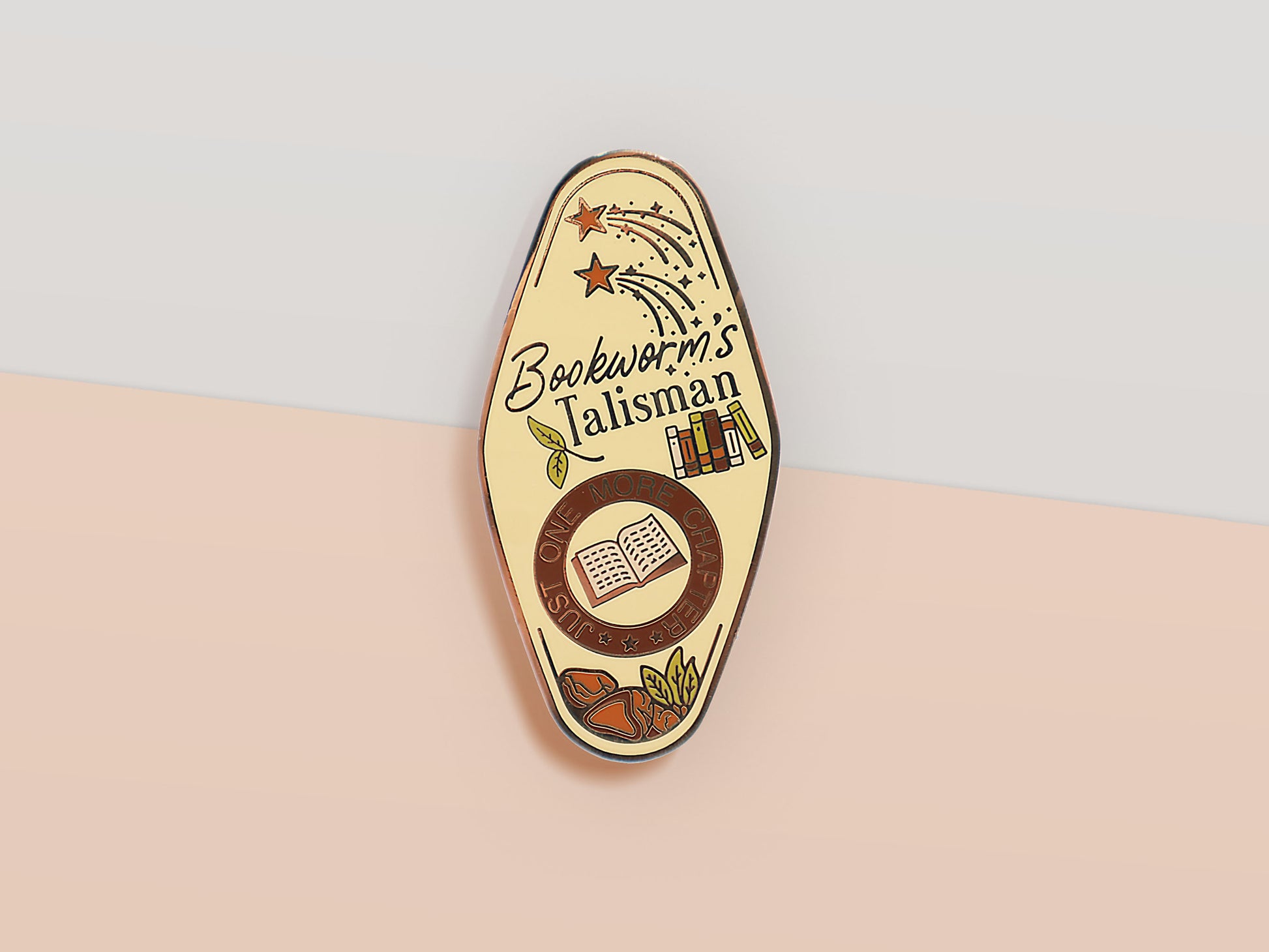 Gold Enamel Talisman Pin with lemon and brown design and the words Bookworm's Talisman, Just One More Chapter. The pins design includes open books and a book shelf, two shooting stars, as well as plants and crystals.