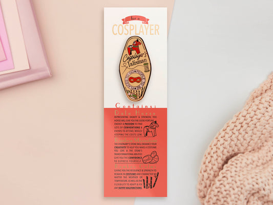 Gold Enamel Talisman Pin with yellow and red design and the words Cosplayer's Talisman sits on a long white and red backing card with gold accents. The backing card has details the symbolism of the different design elements of the Talisman pin.
