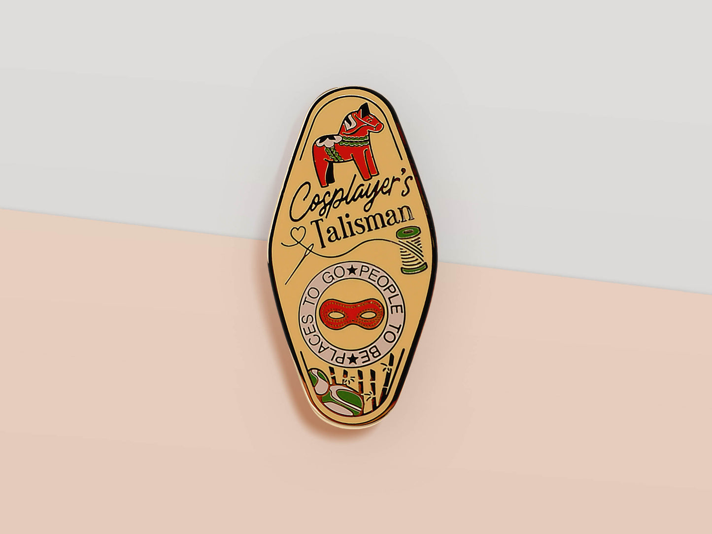 Gold Enamel Talisman Pin with yellow and red design and the words Cosplayer's Talisman, Places To Go People To Be. The pins design includes a mask and needle and thread, a wooden horse, as well as plants and crystals.