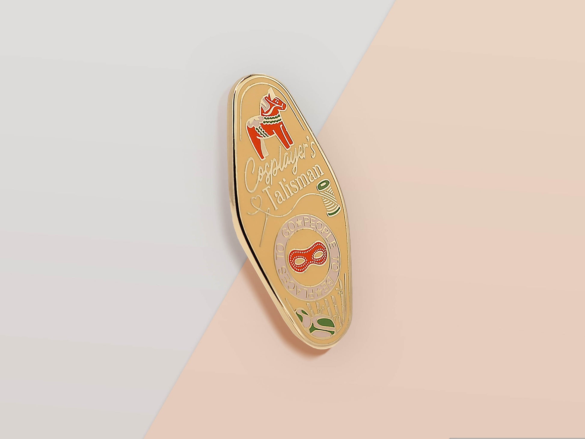 Gold Enamel Talisman Pin with yellow and red design and the words Cosplayer's Talisman, Places To Go People To Be. The pins design includes a mask and needle and thread, a wooden horse, as well as plants and crystals.