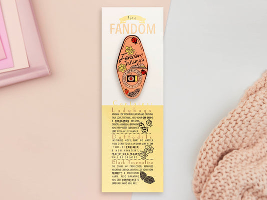 Gold Enamel Talisman Pin with pink design and the words Fandom Talisman sits on a long white and yellow backing card with gold accents. The backing card has details the symbolism of the different design elements of the Talisman pin.