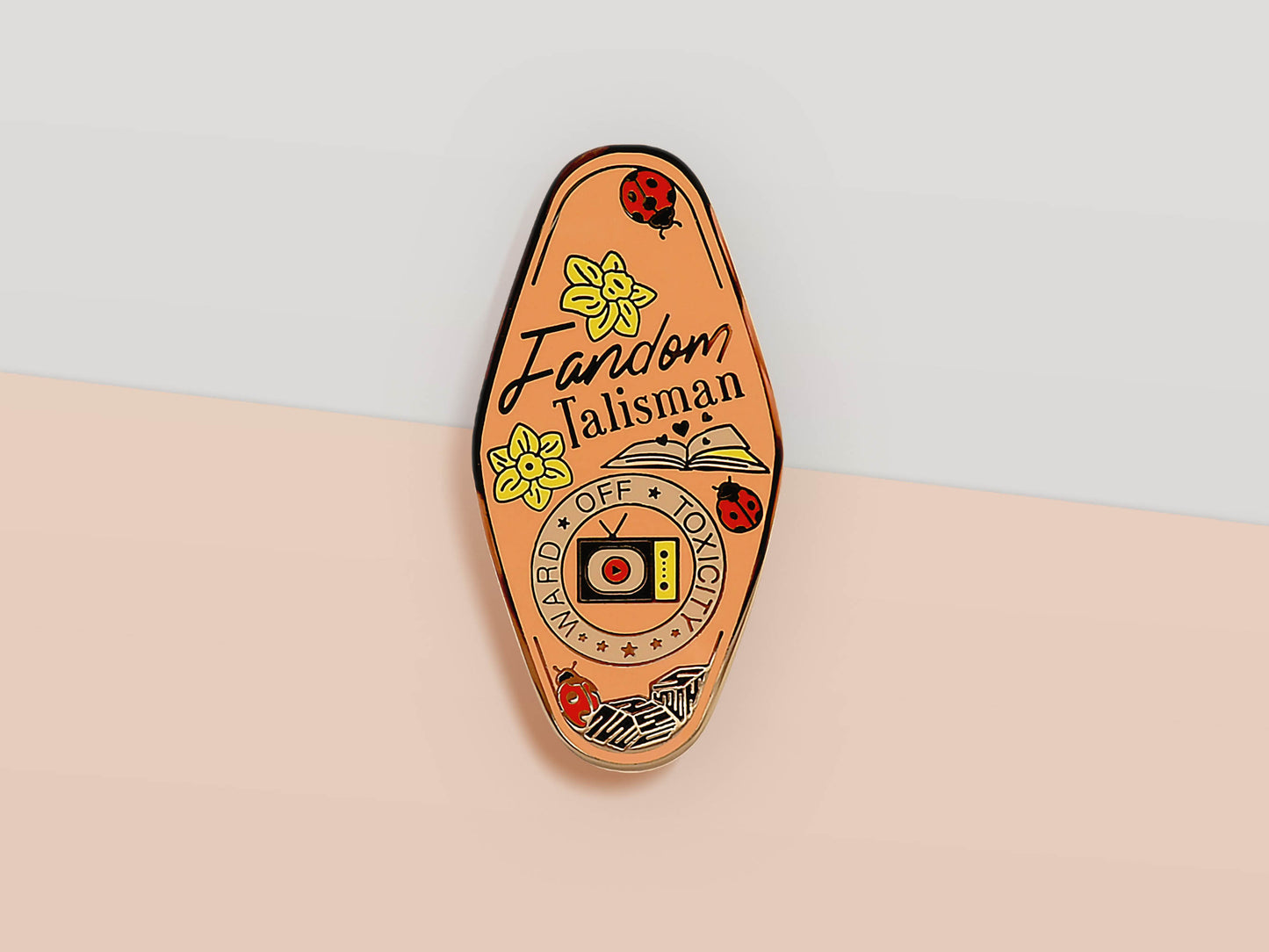 Gold Enamel Talisman Pin with pink design and the words Fandom Talisman, Ward of toxicity block. The pins design includes a television and an open book, ladybugs, as well as flowers and crystals.