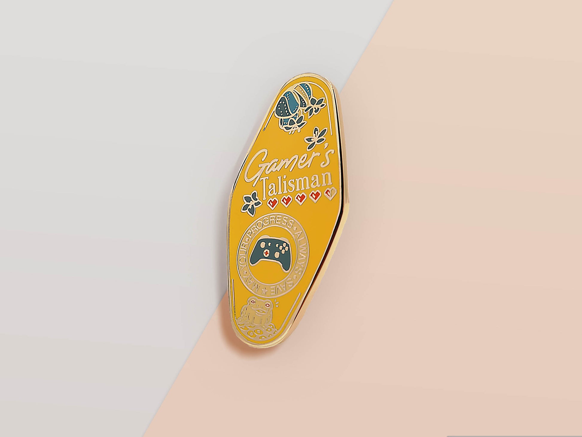Gold Enamel Talisman Pin with yellow design and the words Gamer's Talisman, May Your Progress Always Save. The pins design includes a game console controller and health hearts, A jin Chan money toad, as well as plants and crystals.