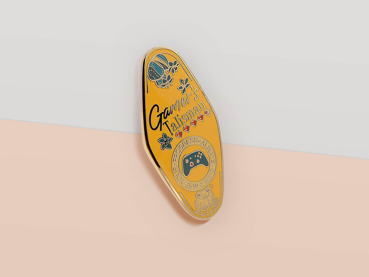 Gold Enamel Talisman Pin with yellow design and the words Gamer's Talisman, May Your Progress Always Save. The pins design includes a game console controller and health hearts, A jin Chan money toad, as well as plants and crystals.