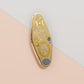 Gold Enamel Talisman Pin with yellow design and the words Mother's Talisman, Wisdom Love Strength. The pins design includes a a mother and child's hand, the three mothers moons, as well as flowers and crystals.