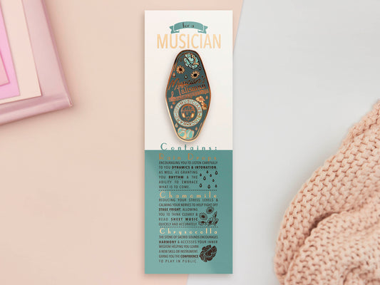 Gold Enamel Talisman Pin with blue design and the words Musician's Talisman sits on a long white and blue backing card with gold accents. The backing card has details the symbolism of the different design elements of the Talisman pin.