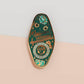 Gold Enamel Talisman Pin with blue design and the words Musician's Talisman, Ward of Musician's block. The pins design includes headphones and sheet music, raindrops, as well as flowers and crystals.