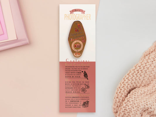 Gold Enamel Talisman Pin with brown design and the words Photographer's Talisman sits on a long white and red backing card with gold accents. The backing card has details the symbolism of the different design elements of the Talisman pin.