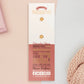 The back of the long white and red backing card with gold accents. The backing card details the symbolism of the different design elements of the Photographer's Talisman pin. Also shown is the two gold locking pin backs attached to the card.