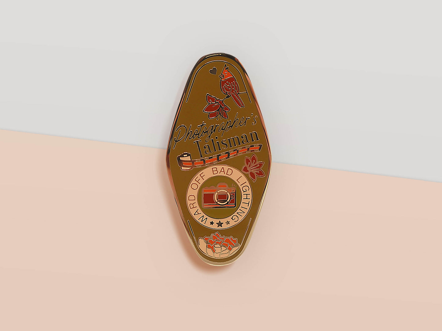 Gold Enamel Talisman Pin with brown design and the words Photographer's Talisman, Ward of bad lighting. The pins design includes a camera and a film, a red bird, as well as flowers and crystals.