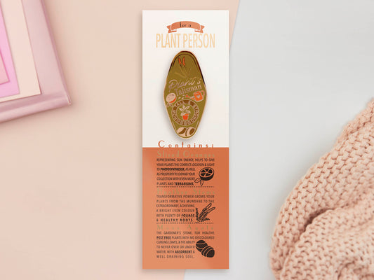 Gold Enamel Talisman Pin with green design and the words Plant's Talisman sits on a long white and orange backing card with gold accents. The backing card has details the symbolism of the different design elements of the Talisman pin.