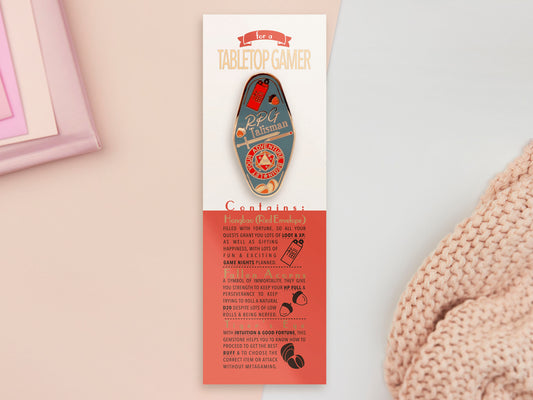 Gold Enamel Talisman Pin with red and blue design and the words RPG Talisman sits on a long white and red backing card with gold accents. The backing card has details the symbolism of the different design elements of the Talisman pin.