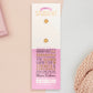The back of the long white and pink backing card with gold accents. The backing card details the symbolism of the different design elements of the Student's Talisman pin. Also shown is the two gold locking pin backs attached to the card.