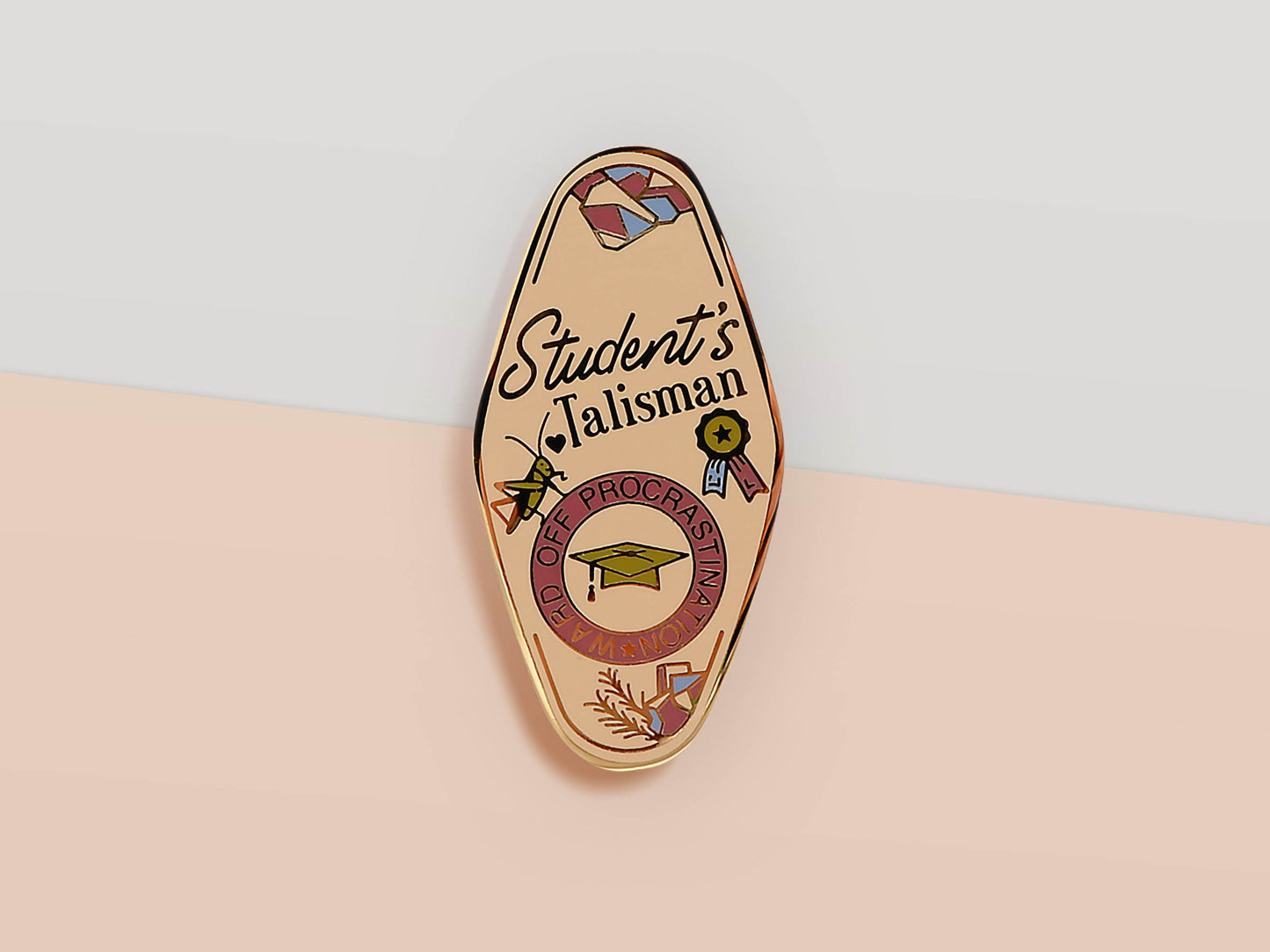 Gold Enamel Talisman Pin with white design and the words Student's Talisman, Ward of procrastination. The pins design includes a graduation cap and a rosette, a grass hopper, as well as plants and crystals.