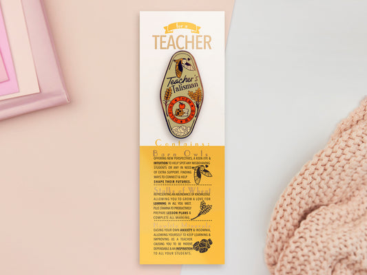 Gold Enamel Talisman Pin with green design and the words Teacher's Talisman sits on a long white and yellow backing card with gold accents. The backing card has details the symbolism of the different design elements of the Talisman pin.