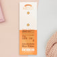 The back of the long white and orange backing card with gold accents. The backing card details the symbolism of the different design elements of the Artist's Talisman pin. Also shown is the two gold locking pin backs attached to the card.