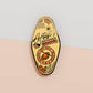 Gold Enamel Talisman Pin with yellow design and the words Artist's Talisman, Ward of Artist's block. The pins design includes a paintbrush, pencil, computer stylus and paint, spider's webs, as well as flowers and crystals.