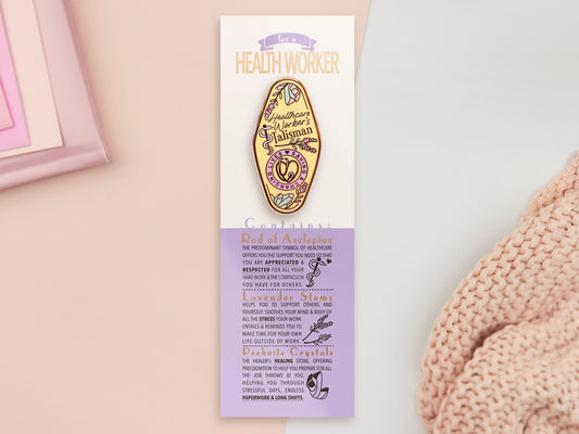 A Health Worker's Talisman gold enamel pin on a gold foiled backing card that outlines the symbolism of the talisman including information and imagery on the Rod of Asclepius, Lavender Stems and Prehnite Crystals