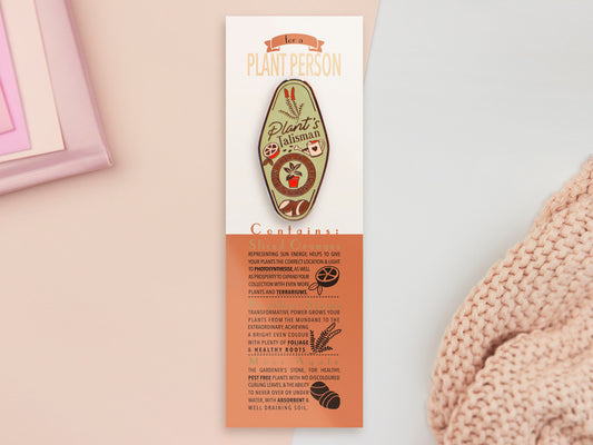 A Plant Person's Talisman gold enamel pin on a gold foiled backing card that outlines the symbolism of the talisman including information and imagery on sliced oranges, heather stems, and moss agate
