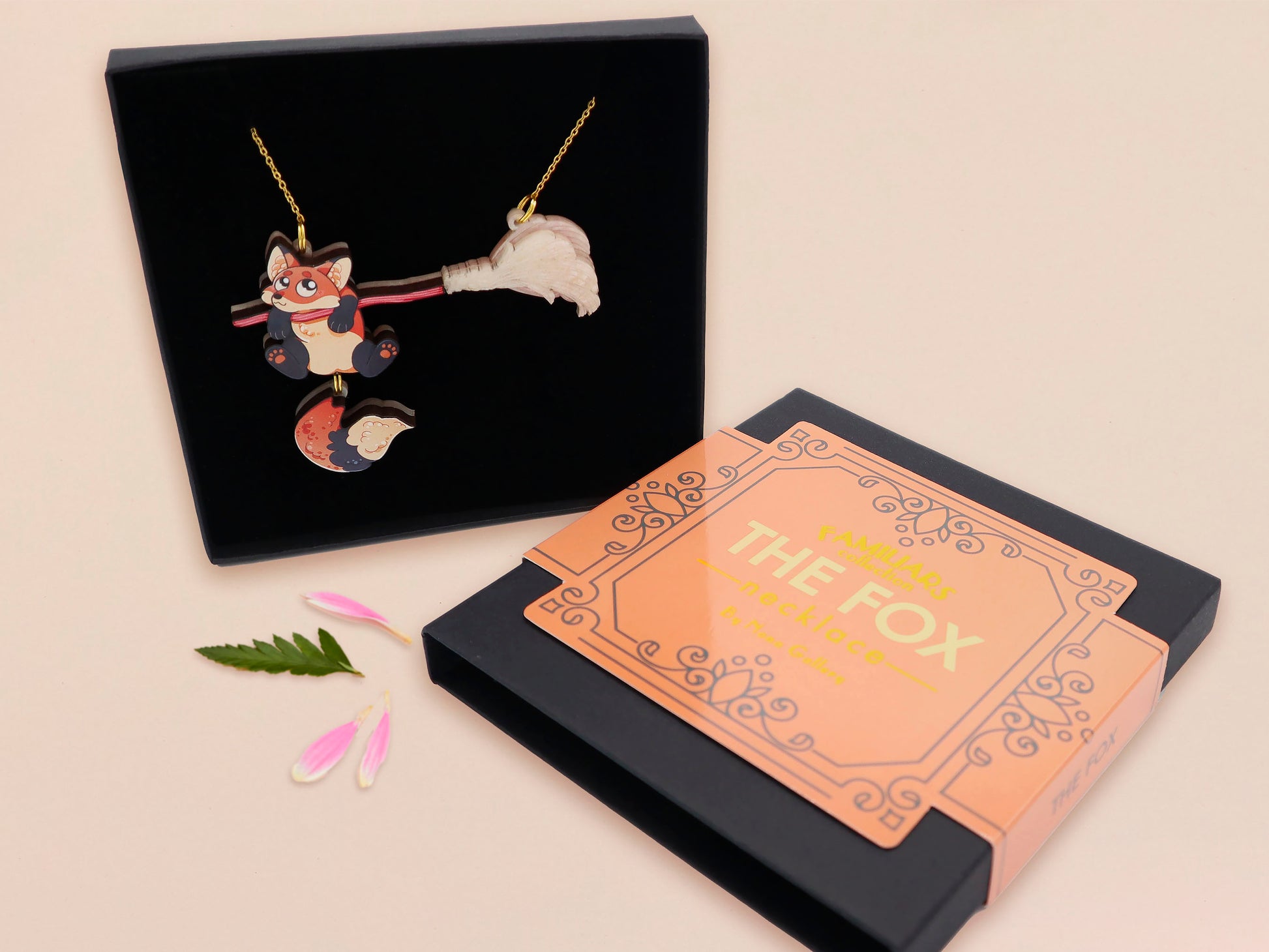 Mixed material handmade necklace of chibi cartoon Fox hanging on to a pearlescent witches broomstick, with a gold chain and black gift box with an orange familiars collection gift sleeve.