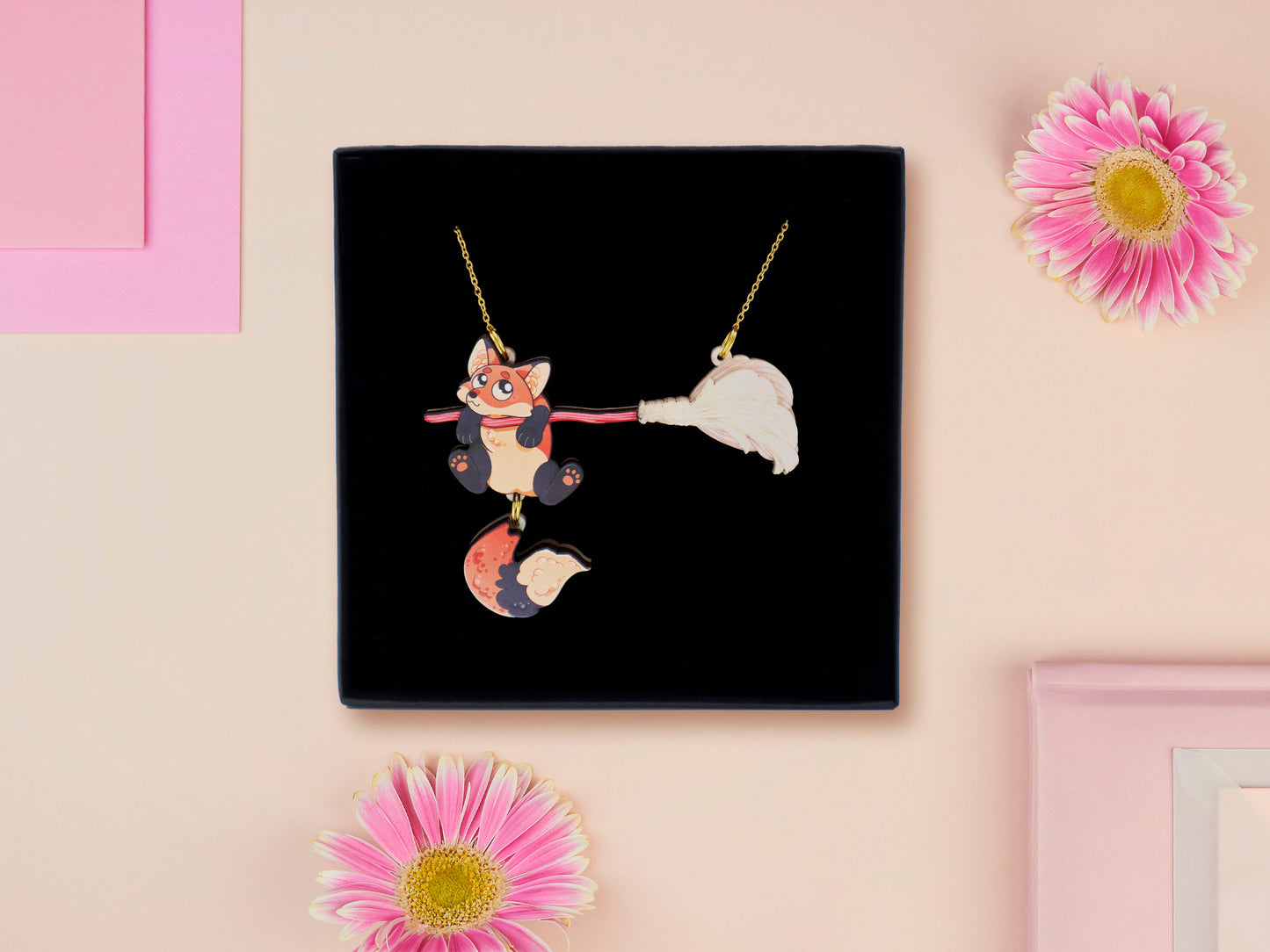 Mixed material handmade necklace of chibi cartoon Fox hanging on to a pearlescent witches broomstick, with a gold chain and black gift box.