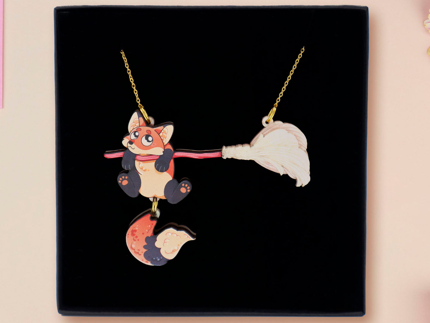 Mixed material handmade necklace of chibi cartoon Fox hanging on to a pearlescent witches broomstick, with a gold chain and black gift box.